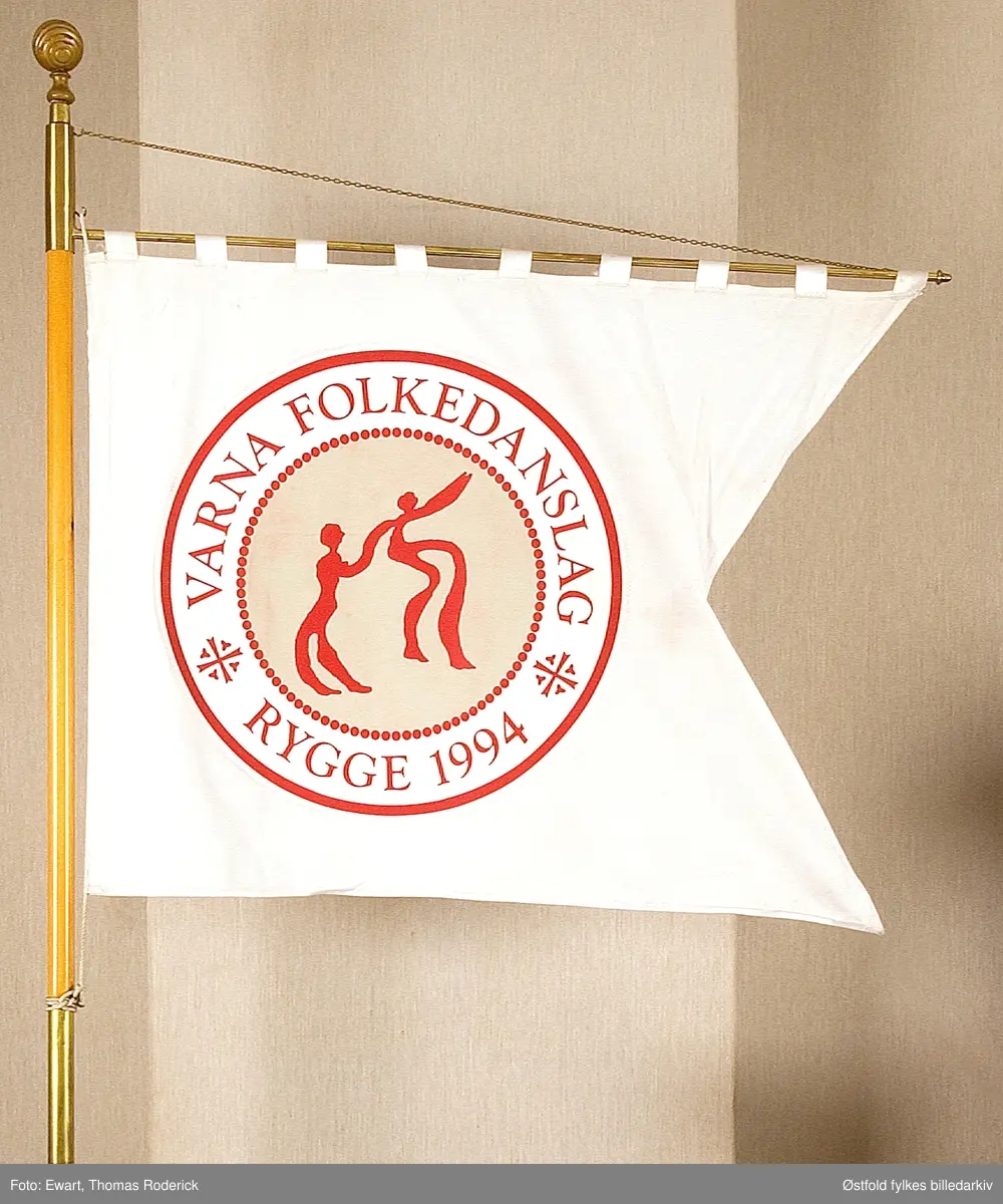 Splittbanner til Varna folkedanslag, Rygge skipa 12. 01. 1994, tilslutta NU / Eidsiva Fylkeslag.. Forside. 
Splittbanner av bomullschins 100 x 180 cm fra 1998, silktrykk med applikert emblem som er likt på begge sider. 
Motiv: eit dansande par som er kopi av helleristning frå bronsealderen på Kalnes i Tune, Østfold. Perlering og johanitterkrossa frå Værne kloster i Rygge. Den ytste ringen symboliserear dei yngste medlemmane av laget, perleringen innanfor symboliserar barna som blir verna av dei vaksne. Johanitterkrossa og perleringen er frå seglet til prioren på Værne kloster, som fanns i Rygge frå omlag 1190 til om lag 1570.  Perleringen og krossa symboliserar også på sin måte at laget høyrer til den kristne kulturarven og til Rygge. Banneret er laga av eigen komite i laget og av Finn Solvang AS, Oslo. Utføringa er gjort hjå Reklametrykk i Moss ved Terje Lindholm.