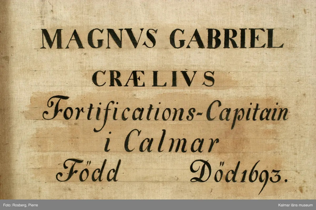 Porträtt av Magnus Gabriel Craelius, Fortifications-Capitain i Kalmar. Grön livrock av polskt snitt. Skinnuppslag på ärmarna. Stora ficklock med många knappar. Värja och gehäng. Spetskrås vid hals mm. Peruk. I högra handen käpp och handske, på vänstra handen älghudshandske.