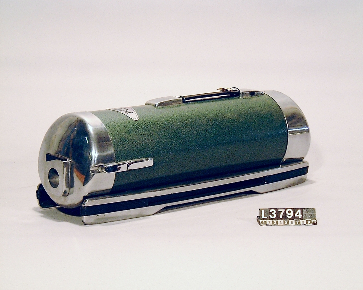 Cylinderdammsugare i grön metallic med fram och bakstycke i blank metall. Medar i krom med hjul under. Kabel, rör, munstycken och vätskespridare finns. Apparaten med tillbehör ligger i en koffert/väska. Slangen saknas.