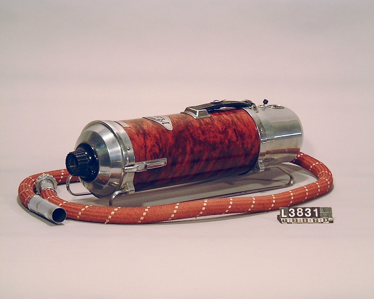 Cylinderdammsugare klädd i vinröd/flammig pegamoid. Fram och bakstycke samt medarna är i metall. Apparaten ligger i en koffert/väska. Under apparaten finns en etikett med Nr 31. Apparaten är alldrig använd, flera av tillbehören är inslagna i papper. Alla delar finns.