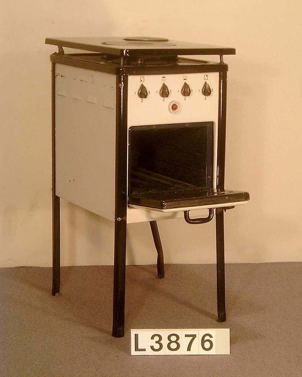 Golvspis på fyra st. svarta emaljerade ben. Två kokplattor, uppfällbar överhäll som är svartemaljerat. Frampartiet och sidorna är vitemaljerade. Ugn, men inget värmeskåp. Strömställarna har tre lägen. Ugnen har ingen termostat, utan två separata vred ett för över och ett för undervärme.