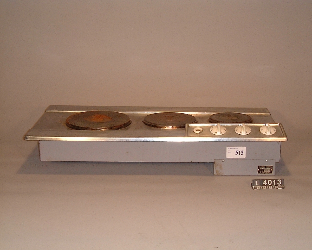 Restauranghäll i rostfritt stål med tre plattor på rad. Längst ned i högra hörnae på hällen  sitter vreden upphöjd plint. Vreden är i vit plast.