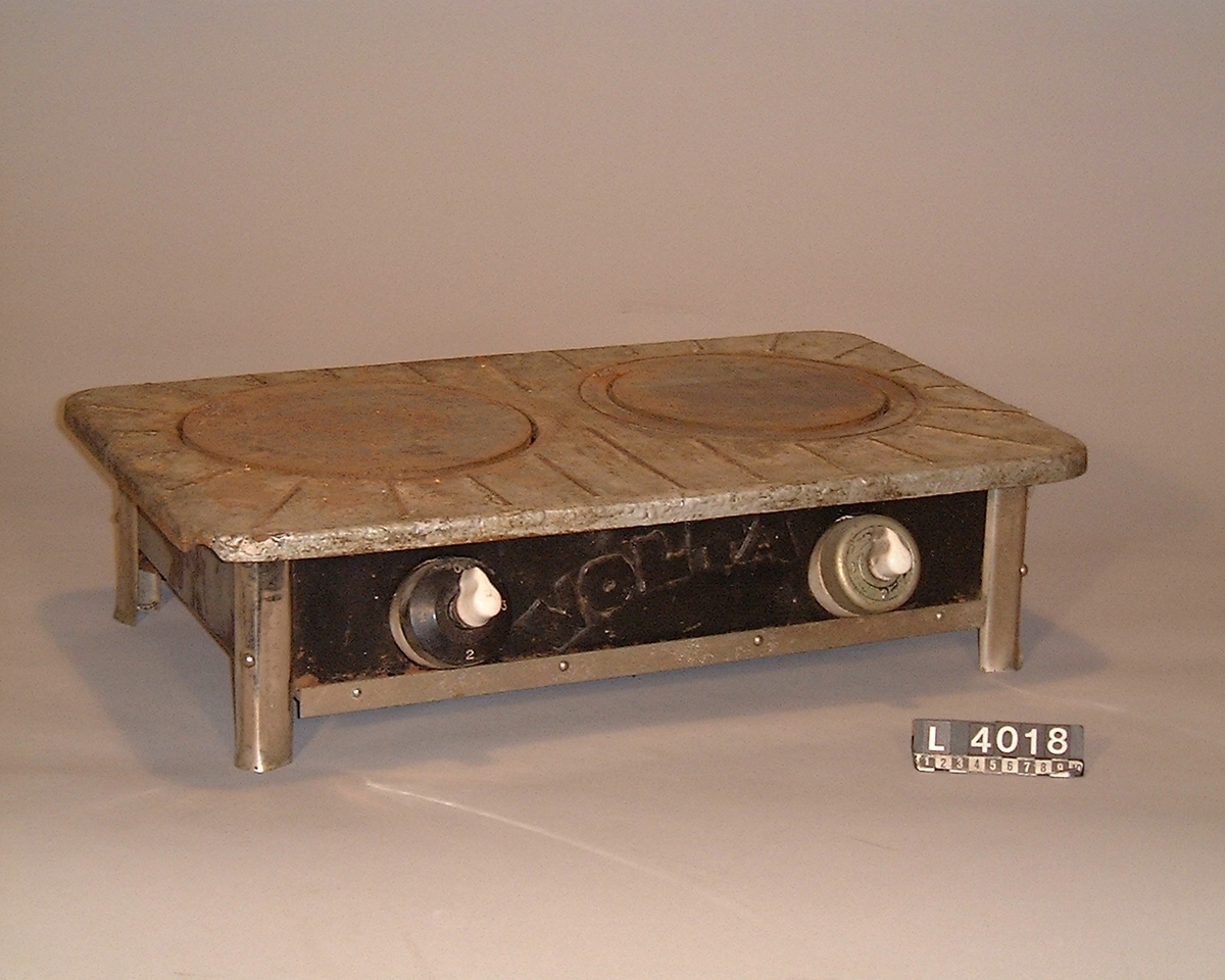 Bänkspis med två plattor. På fronten som är av svartmålad plåt sitter två strömbrytare i porslin med kåpor av blank metall. I varje hörn finns kromade ben, resten är svart utom hällen som är målad i blank metall.