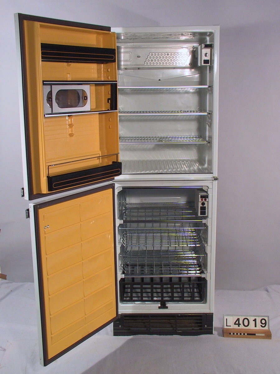 Kombinationsskåp Elektro-Helios med dubbla kompressorer, ett för kyldelen och ett för frysdelen. Kylskåpet är vitt med handtag inspirerade från bilindustrin. Inredningen är vit och dörr- insidorna går i gult.