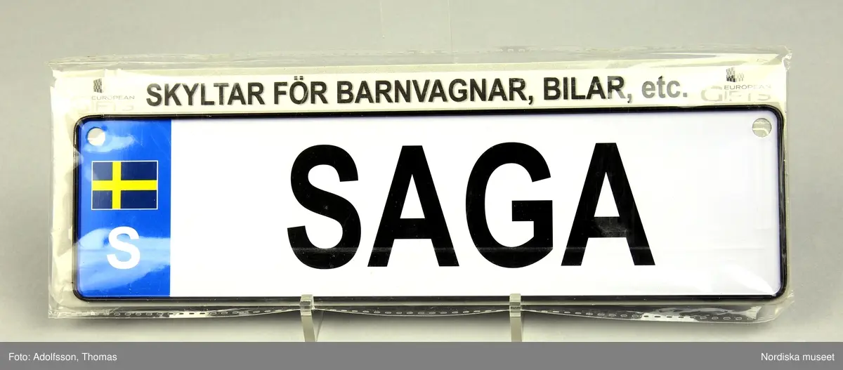 Namnskylt av plåt, avsedd för barnvagn i originalförpackning. Är utformad som en registreringsskylt för bil. Rektangulär, vit med svart ram. På skylten finns namnet Saga samt en svensk flagga och nationsbokstaven S. I övre hörnen hål.

/ C Hammarlund-Larsson 2019-01-25