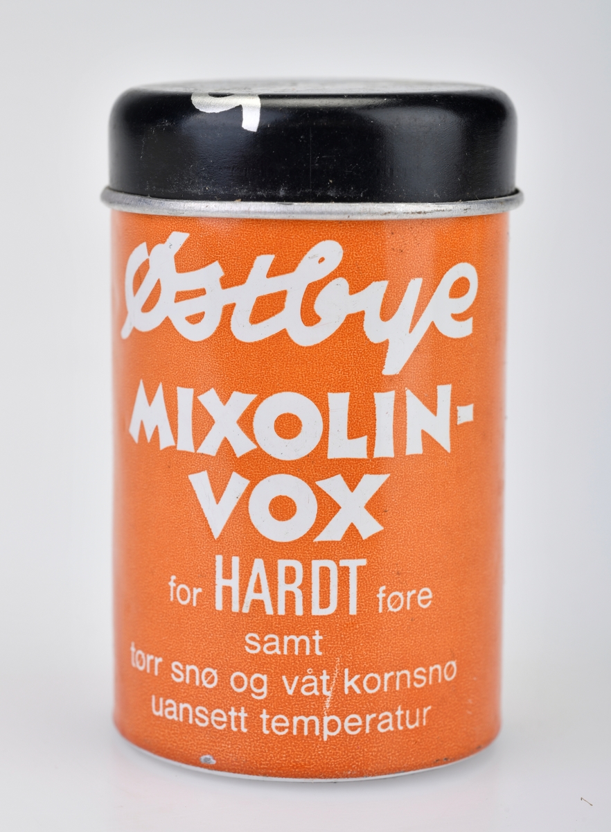 En boks/sylinder med skismurning av typen Østbye Mixolinvox for hardt føre, tørr sne og våt kornsne. Boks og lokk er av metall. Metallet skal rives av etter hvert som det brukes av skismurningen. Boksen er oransje med hvit skrift, lokket er svart med sølvfarget skrift. For tekst på for- og bakside - se "Påført tekst/merker".  Det er informasjon om bruk på engelsk, tysk og norsk. Voksen er oransje på farge.