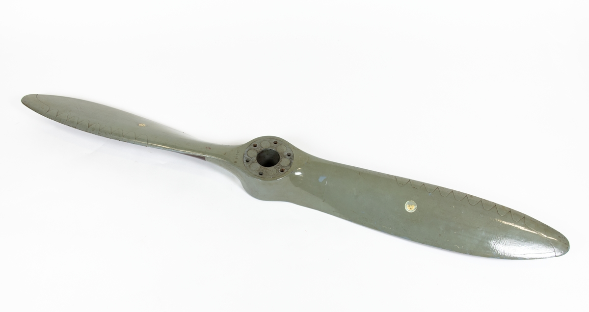 Tvåbladig träpropeller till SK 12. Propellern är målad grå och har skoning på bladen. Baksidan av propellerbladen är bruna. 
Propellern ligger i en trälåda. På locket står det att den tillhör SK 12/70.