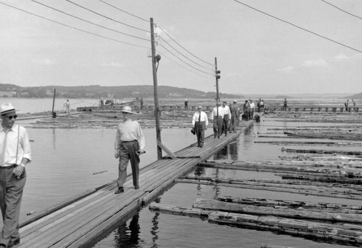 Fra direksjonsbefaring i Glomma fellesfløtingsforening sommeren 1958.  Fotografiet viser ei rekke av menn i lyse skjorter som kom vandrende på en av lensas flåteganger.  Til høyre for karene ligger en del barket fløtingstømmer og flyter i vannskorpa på ei av ”rennene” (passasjene) i lenseanlegget.  Fotografiet er antakelig fra den øvre delen av sorteringsanlegget, der hvor tømmeret ble sluppet inn fra den ovenforliggende beholdningslensa.  Direksjonen besto av representanter for trelast- og treforedlingsbedriftene som kjøpte virke langs Glommavassdraget, og av representanter for skogeierne i nedslagsfeltet.  Også sentrale medarbeidere i administrasjonen og funksjonærer fra de anleggene og distriktene som fikk besøk, pleide å være med på befaringene.