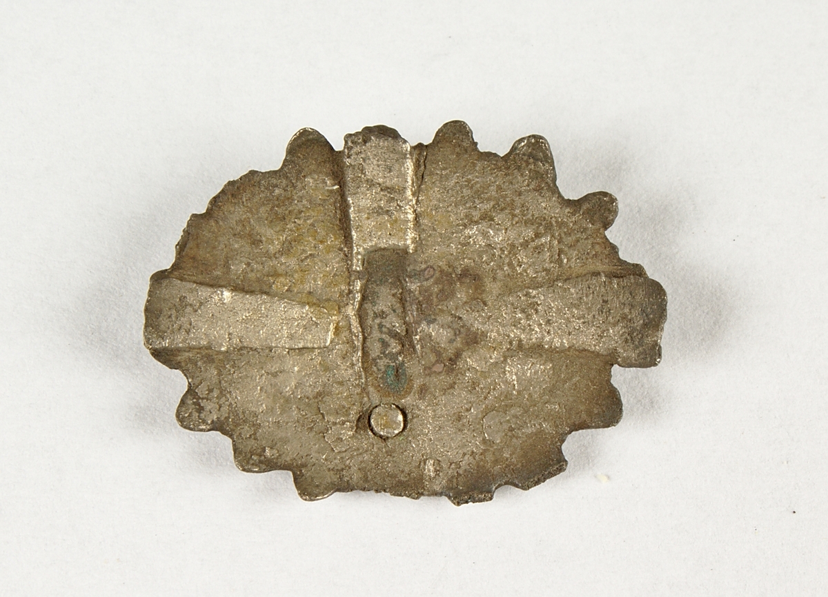 Ovalt smycke i silver med infattning för sten. Stenen är dock borta. Kanterna runt smycket är kantade.