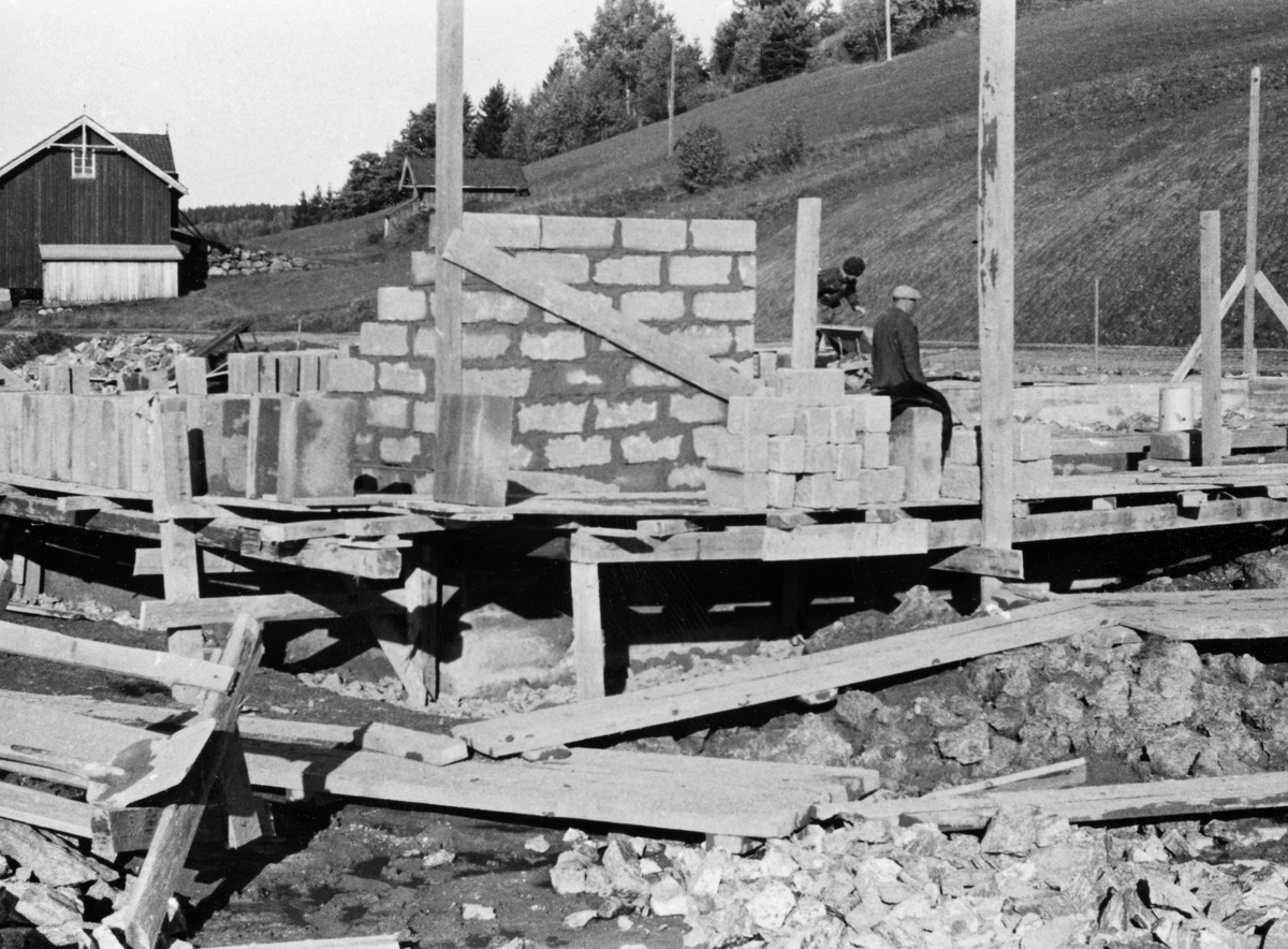 Muring av nytt verkstedbygg ved Glomma fellesfløtingsforenings anlegg på Glennetangen i Varteig under bygging høsten 1953.  Fotografiet er tatt sørfra, der det er murt et hjørne.  Bildet viser hvordan det var reist en plattform, et lavt stillas, av tre på utsida av bygningen.  På denne plattformen lå det, da dette fotografiet ble tatt, en del såkalte «tretong-blokker», som ble brukt under murerarbeidet.  Til høyre for det påbegynte hjørnet, men noe lengre inne i bildet, ser vi to arbeidere i virksomhet.  Bakenfor anleggsområdet skimter vi noe av bilvegen forbi Furuholmen, og noe av jordvegen på bruket.  Helt til venstre i bildet ser vi sørgavlen på Furuholm-låven. 
 
Bygginga av det nye verkstedet var ett av flere tiltak som ble gjort som forberedelse til at en rekke funksjoner skulle flyttes fra det gamle fløtingssenteret ved Nes til Furuholmen.  Dette ble gjort fordi Furuholmen lå like ved Glennetangen, der hovedlensa for denne delen av vassdraget hadde ligget siden siste halvdel av 1930-åra.  Flyttinga av så vel funksjonærer som tekniske servicefunksjoner ble sluttført i 1958-59.