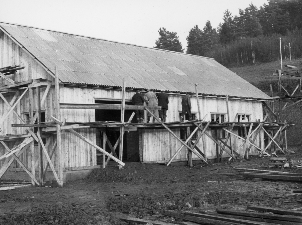 Den nye verkstedbygningen ved Glomma fellesfløtingsforenings anlegg på Furuholmen i Varteig, fotografert idet byggearbeidet begynte å gå mot slutten seinhøstes (i november) 1953.  Det dreide seg om en forholdsvis høy enetasjes konstruksjon med eternittplatetekket saltak.  Bygningen skulle i hovedsak brukes til metallarbeider (sveising m. m. ), og den ble derfor murt.  Utvendig valgte man imidlertid, i stedet for å pusse murene, å kle bygningen med et trepanel.  Dette arbeidet skal ha vært i en avsluttende fase da fotografiet ble tatt.  Fire menn sto på et stillas i en høyde omtrent midt på langveggen, like ved portåpningen.  På gavlveggen (til venstre i bildet) var det stillas i to høyder.  

Bygginga av det nye verkstedet var ett av flere tiltak som ble gjort som forberedelse til at en rekke funksjoner skulle flyttes fra det gamle fløtingssenteret ved Nes til Furuholmen.  Dette ble gjort fordi Furuholmen lå like ved Glennetangen, der hovedlensa for denne delen av vassdraget hadde ligget siden siste halvdel av 1930-åra.  Flyttinga av så vel funksjonærer som tekniske servicefunksjoner ble sluttført i 1958-59. 
