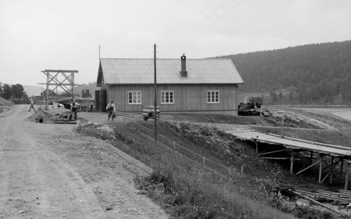 Glomma fellesfløtingsforenings verkstedbygning på Furuholmen i Varteig, fotografert sommeren 1954, et snaut år etter at bygningen var ferdig.  Fotografiet er tatt fra bilvegen nord for anlegget, og vi ser den nordre langveggen med tre vindusåpninger og med skorsteinspipe.  Fra bakkekammen nord for bygningen var det bygd ei trebru (på stolper) ned mot vassdraget og slippen for fløtingsbåtene.  Denne brua ble brukt i samband med uttak av dampmaskiner og innsetting av dieselmotorer i slepebåter.  Til høyre for verkstedbygningen sto det et «tårn», bygd av trestolper, som ble brukt til heising av tunge objekter som skulle ned fra eller opp på lasteplan.  Foran det nevnte tårnet lå det en diger sandhaug, der det sto en mann og spadde da fotografiet ble tatt. 

Reisinga av nye verkstedbygninger, våningshus og kaianlegg og flere andre byggeprosjekter ved Furuholmen var ledd i et arbeid for å kunne flytte virksomhet fra det gamle fløtingssenteret ved Nes, cirka to mil høyere opp i vassdraget, til Furuholmen.  Dette ble gjort fordi Furuholmen lå like ved Glennetangen, der hovedlensa for denne delen av vassdraget hadde ligget siden siste halvdel av 1930-åra.  Flyttinga av så vel funksjonærer som tekniske servicefunksjoner ble sluttført i 1958-59.