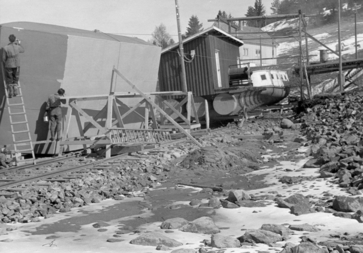 Vaierpram av jern, reist på høykant ved Glomma fellesfløtingsforenings bryggeanlegg på Furuholmen i Varteig i Østfold.  Dette var vedlikeholdsarbeid.  Prammen ble trukket opp ved hjelp av en traktor med firehjulsdrift, og jekket opp slik at den ble stående på høykant.  Det siste skal det ha vært Arnt Svartedal d. y. som fant på.  I denne stillingen ble prammen først sandblåst, deretter malt.  Dette fotografiet ble tatt mens malingsarbeidet pågikk.  Vi ser undersida av farkosten, to malere og tre bukker med en gangbane, antakelig oppsatt slik at malerne skulle nå helt opp.  Til høyre for prammen ser vi ei brakke i bordkledd bindingsverk med pulttak, og med underliggende flytelegemer av tomme jernfat.  Bak den igjen lå båten Pål, en varpebåt som var ombygd med tanke på transport av mannskaper til og fra lensene.  Mens prammen, brakka og båten ligger på rekke i strandsonen, skimter vi høyere oppe i bakken formannsboligen ved anlegget.  Den pramtypen som er hovedmotiv på dette fotografiet ble, som betegnelsen «vaierpram» indikerer, brukt til å transportere bind til tømmermoser.  Prammene var så robuste og stødige at de også ble brukt til transport av tunge objekter som det ikke var så enkelt å få lastet på eller losset fra de andre båttypene fløterne disponerte.