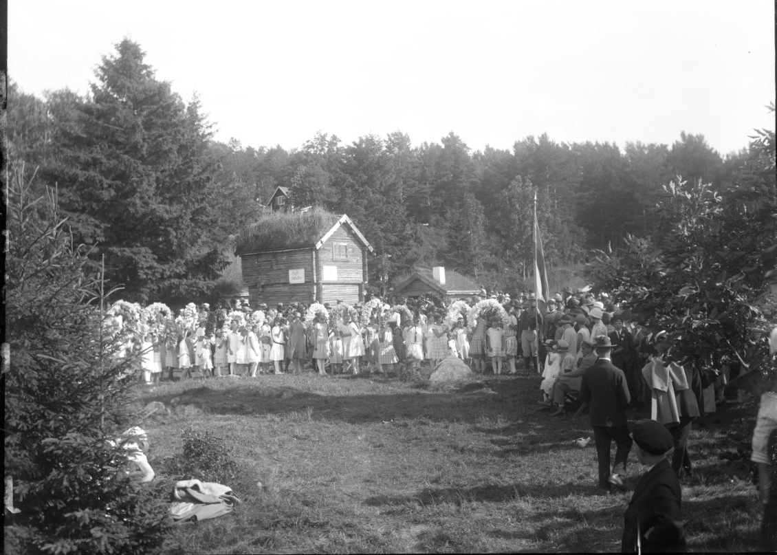 Folksamling, barn och vuxna vid Friluftsmuseet på Grännaberget, det är Hembygdsfest. Barnen håller blomsterbågar i händerna. De står framför pappersmakarbostället vars loftbod har en skylt: "Kaffebiljetter".