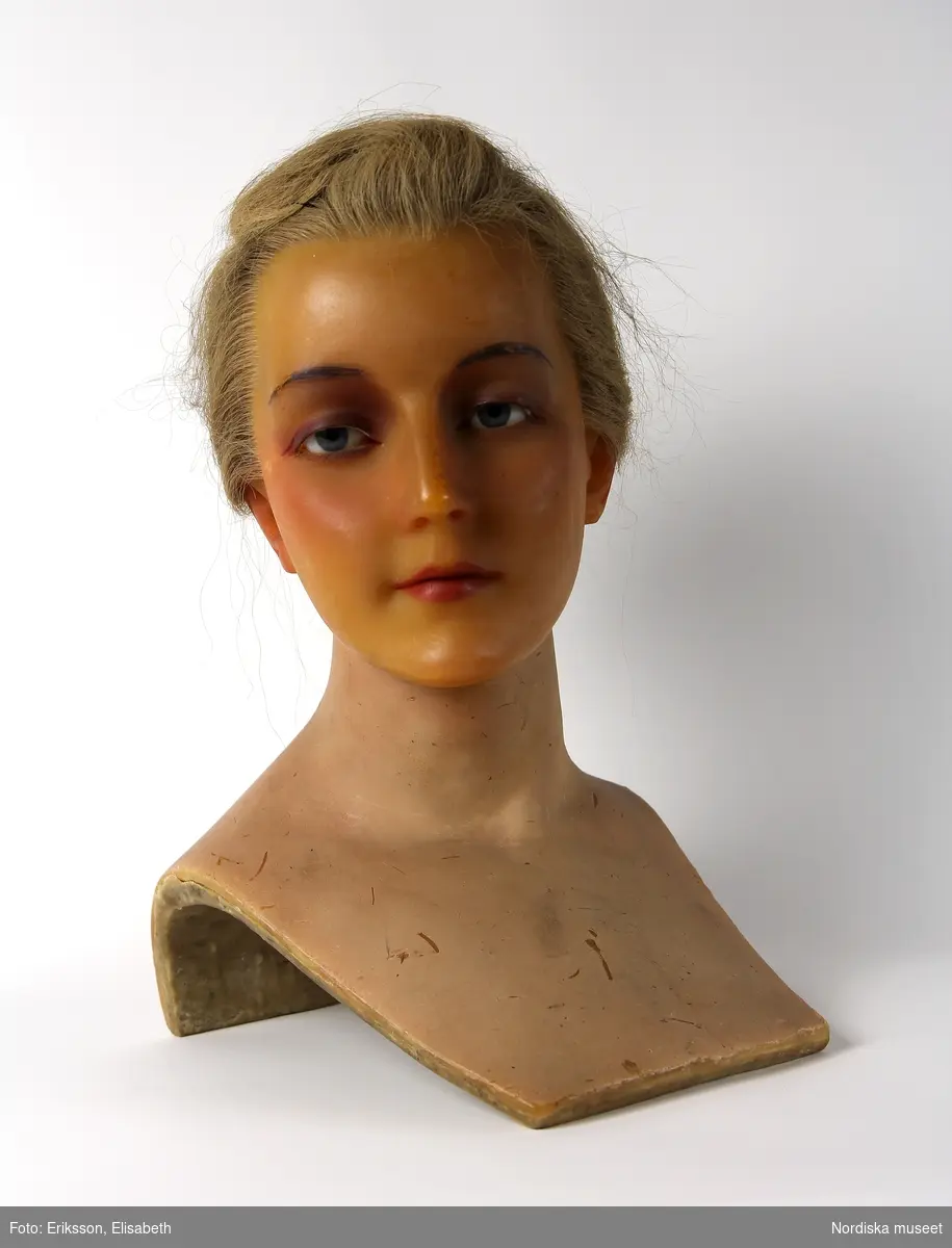 Skyltdockshuvud, kvinna, av vax med riktigt hår, ögonfransar och ögon av glas. Med hals och övre del av torso. På baksidan av torso text "Paul Baschwitz / Berlin C". Blont hår friserat i knut (hårnålar ingår ej i förvärvet).
Anm. spricka vid höger och vänster öga och vax gulnat.
/Helen Persson 2018-02-13