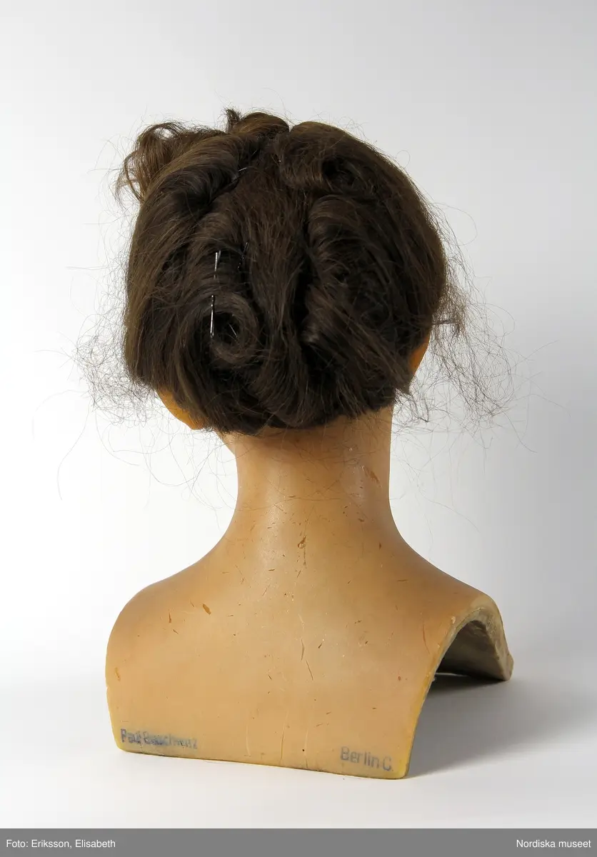 Skyltdockshuvud, kvinna, av vax med riktigt hår, ögonfransar och ögon av glas. Med hals och övre del av torso. På baksidan av torso text "Paul Baschwitz / Berlin C". Håret friserat i 1940-tals frisyr (hårnålar ingår ej i förvärvet).
Anm. spricka vid vänster öga och vax gulnat.
/Helen Persson 2018-02-13