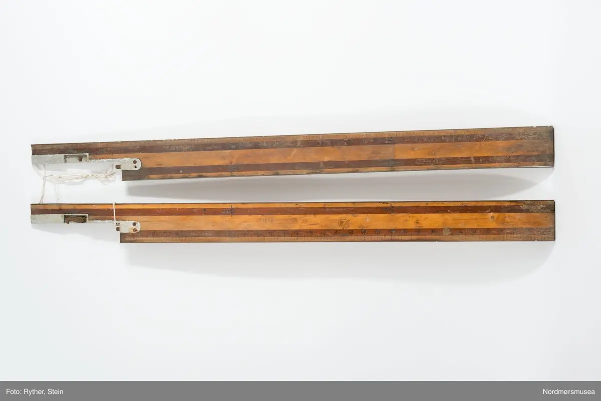 Linjalen er på 72 inches = 6 feet. Oppmerkningen slått inn med stempler. Delt ved 32 1/2" - 39 1/2". Her er satt på et aluminiumsbeslag emd tappesystem og krok. Tallene er stemplet på ei stripe av brunt tre, resten er lyst treverk. Messingskinner skrudd til over- og undersiden av linjalen.