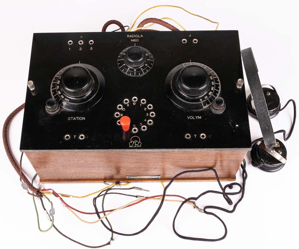 Batteridriven 2 rörs radioapparat. Lådan av svart och brunt trä. 2 par hörlurar. Diverse sladdar. På lådans sida en metallbricka "SVENSKA RADIOAKTIEBOLAGET STOCKHOLM" På undersidan provsedel med text: "...RADIOLA M60...".