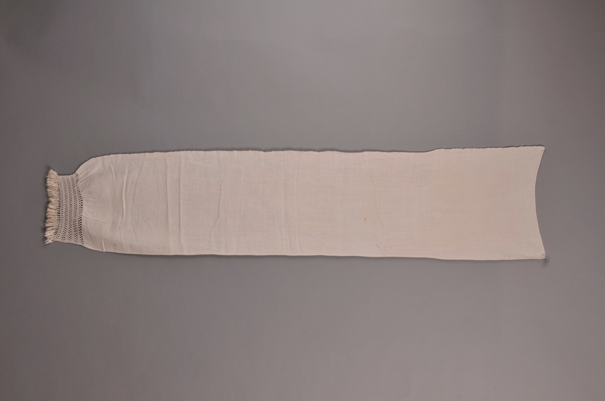 Grautspannklede i kvit, heimvove lin, ca 16 trådar per cm. blonde i firfletting på den eine kortsida. Små duskar i enden av blonda.