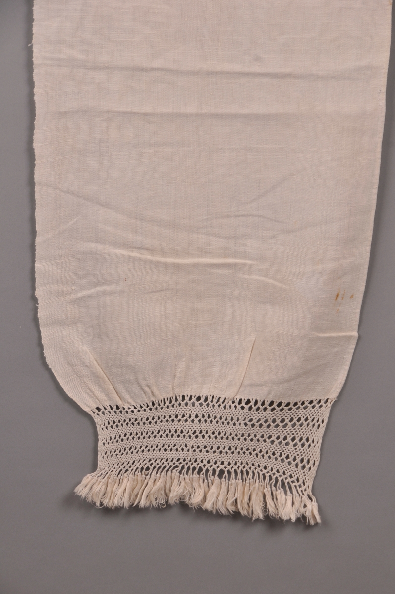 Grautspannklede i kvit, heimvove lin, ca 16 trådar per cm. blonde i firfletting på den eine kortsida. Små duskar i enden av blonda.