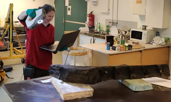 Arkeolog holder en skanner i høyre hånd og en bærbar datamaskin i venstre hånd, og peker skanneren mot en båtdel som ligger på et bord.