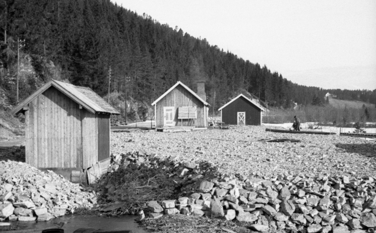 Grunnarbeid med sikte på bygging av båthus for Glomma Fellesfløtingsforenings båter sør for Åsheim i nordenden av Storsjøen i Rendalen.  Dette ble gjort ved å hente stein fra et brudd på vestsida av bygdevegen (nåværende Riksveg 3), over på østsida av vegen, mot sjøen. På fyllinga, like ved vegkanten står det tre små bygninger med saltak [den bakerste delvis skjult), alle tilsynelatende utført i bordkledd bindingsverk.  Den nærmste later til å være en dobygning, som etter fargesettinga og taktekkingsmaterialet å dømme nylig er forlenget mot fotografen.  Bakenfor ses gavlene på to litt større bordkledde bygninger, hvorav i hvert fall den nærmeste har murt pipe for ildsted.  Mellom og til øst for bygningene til høyre) er det altså ei steinfylling.  Et par menn i arbeid på denne fyllinga skimtes langt inne i bildet.  Lia til venstre i bildet er stort sett kledd med granskog.  Helt til høyre i bildet skimtes en grasvoll og gavlen på et våningshus. 