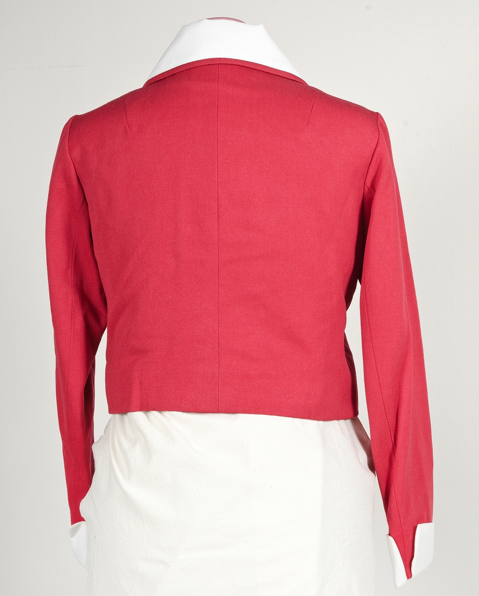 Röd dräktjacka i kypertvävd. Jackan tillhör klänning UM43440a och skärp UM43440c. Jackan är fodrad med rött fodertyg. Knäpps i fram med fyra guldfärgade plastknappar. Utfälld krage. På kragen fastsatt vitt tyg, som ger illusionen av en bluskrage. Vid ärmsluten samma vita tyg som manschetter med sprund.