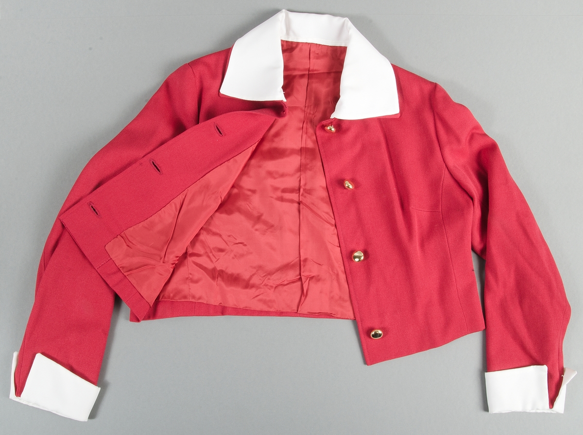 Röd dräktjacka i kypertvävd. Jackan tillhör klänning UM43440a och skärp UM43440c. Jackan är fodrad med rött fodertyg. Knäpps i fram med fyra guldfärgade plastknappar. Utfälld krage. På kragen fastsatt vitt tyg, som ger illusionen av en bluskrage. Vid ärmsluten samma vita tyg som manschetter med sprund.