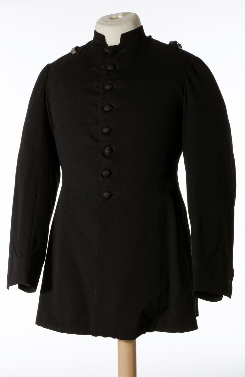 Enkeltspent jakke til uniform i sort ull. Åtte tekstiltrukne knapper i fronten. Kraven er opprettstående og kan lukkes med en metallhekte.  Skuldrene har påsydd hempe på hver side av kragen og hver skulder er dekorert med et 9,5 cm langt vevd, sølvbånd, festet i hver ende vertikalt på skulderen. Baksiden har splitt opp til livlinjen med lommer på hver side inn fra splitten og tre par tekstiltrukne knapper. Nederst på ermene er det utenpåsydd mansjett med spiss og to tekstiltrukne. Innvendig er jakken helforet med sort bomullsfor og vattering i overdelen. Gult fôr i ermene. Opphengshemper i nakken. 

Bukse i mørk blå ull med to lommer bak. Vevd gullbånd i sidene. Gylf lukkes med metallhekte og fire knapper (ant. i tre). Åtte metallknapper utvendig på linning. Hvitt fôr med smale striper i rødt, lyseblått og lysegrått i linning. Sandfarget fôr i lommene.
