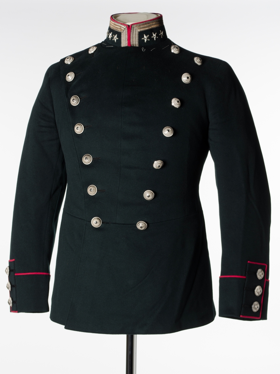 Dobbeltspent jakke til gallauniform i mørk grønn ull, med burgunderrøde detaljer og sølvknapper. Kraven er opprettstående og har ett sølvbånd og tre stjerner. Høyre skulder har to sydde hemper. Fronten har 16 knapper, festet med små plastkanpper på innsiden. Knappene er høye med fargene sølv og hvitt. Knappene har dekorative elementer som henspeiler på et hjul med et horn i midten. Seks små, sydde hemper øverst på venstre side. Baksiden har burgunderrød kantebånd i ryggen, splitt og tre par knapper ved splitten. Splitten er sydd sammen. Innerlomme på venstre side. Nederst på ermene er det tre bånd med knapp og burgunderrøde kantbånd. Innvendig er jakken helforet med sort bomullsstoff og lyst stripete bomullsstoff i ermene. Opphengshemper i ermene. Fronten er vattert. Innvendig belte med tre flate hekter i metall i midjen. Løssnipp er festet på metallnagler. Stivet løssnipp i hvit bomull med fem hull for feste til metallnagler på innsiden av opprettstående krave. Hvite tråklesting i kraven.
Dobbel sølvfarget snor henger på skrå over jakken.