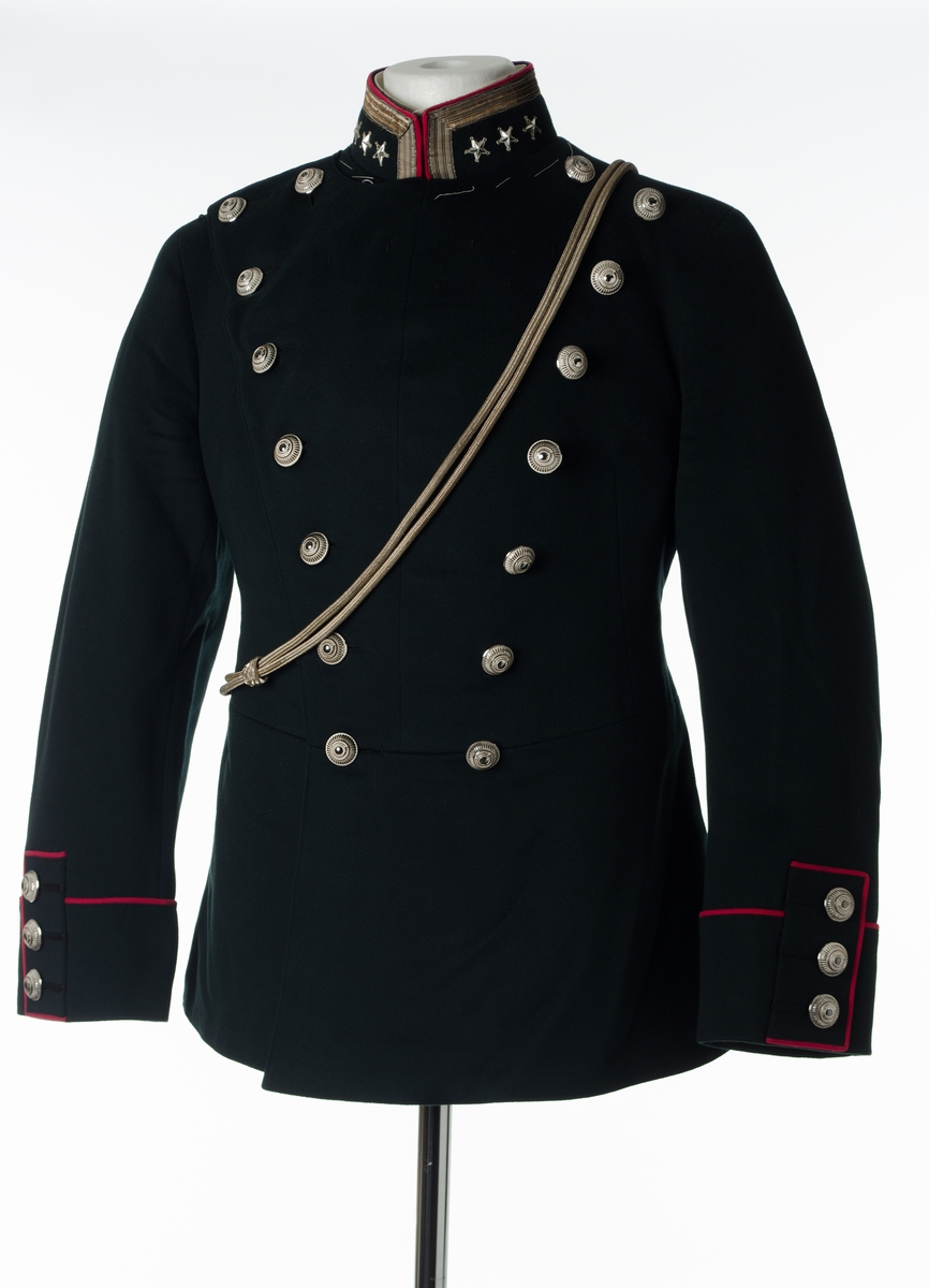 Dobbeltspent jakke til gallauniform i mørk grønn ull, med burgunderrøde detaljer og sølvknapper. Kraven er opprettstående og har ett sølvbånd og tre stjerner. Høyre skulder har to sydde hemper. Fronten har 16 knapper, festet med små plastkanpper på innsiden. Knappene er høye med fargene sølv og hvitt. Knappene har dekorative elementer som henspeiler på et hjul med et horn i midten. Seks små, sydde hemper øverst på venstre side. Baksiden har burgunderrød kantebånd i ryggen, splitt og tre par knapper ved splitten. Splitten er sydd sammen. Innerlomme på venstre side. Nederst på ermene er det tre bånd med knapp og burgunderrøde kantbånd. Innvendig er jakken helforet med sort bomullsstoff og lyst stripete bomullsstoff i ermene. Opphengshemper i ermene. Fronten er vattert. Innvendig belte med tre flate hekter i metall i midjen. Løssnipp er festet på metallnagler. Stivet løssnipp i hvit bomull med fem hull for feste til metallnagler på innsiden av opprettstående krave. Hvite tråklesting i kraven.
Dobbel sølvfarget snor henger på skrå over jakken.