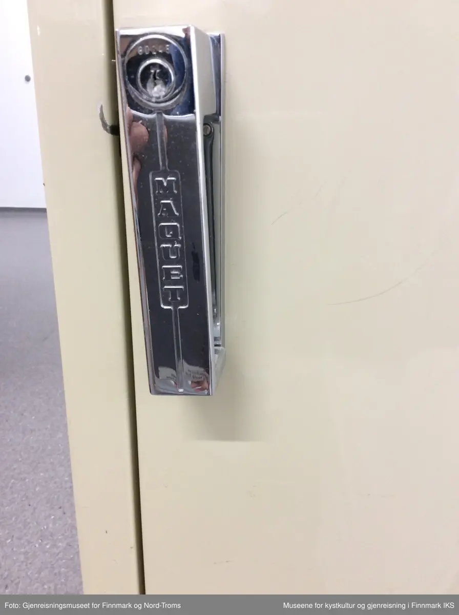 Skapet er av metall og har to dører, ei kvadratisk nede og en rektangulært høy oppe. Begge dører er låsbare og har håndtak av metall som åpnes ved å dreie de til høyre. Det øverste skapet har 5 hyller, den øverste av metall og resten av glass. Skapdøra er av glass med en bøyd nøkkel i låsen. Den nederste skapdøra er av metall og er låst. Hele skapet står på en svart metallsokkel.