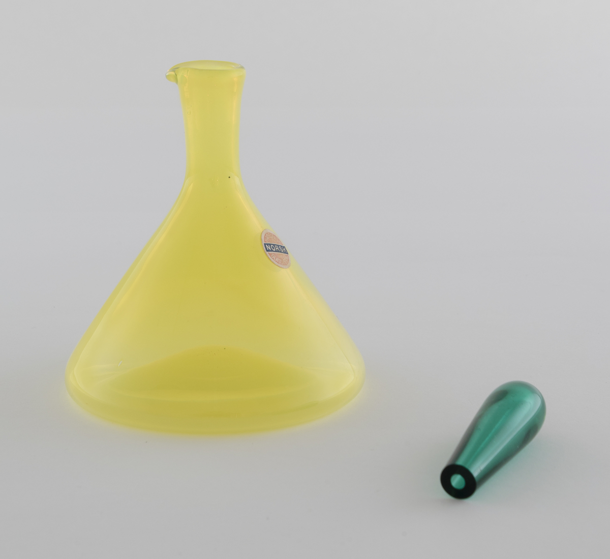 Edikk- eller oljekaraffel med propp i farget glass. Konisk koprus i halvgjennomskinnelig gulfarget glass, som avsluttes i en smal hals med tut. Den dråpeformede proppen er utført i gjennomskinnelig grønnfarget glass.