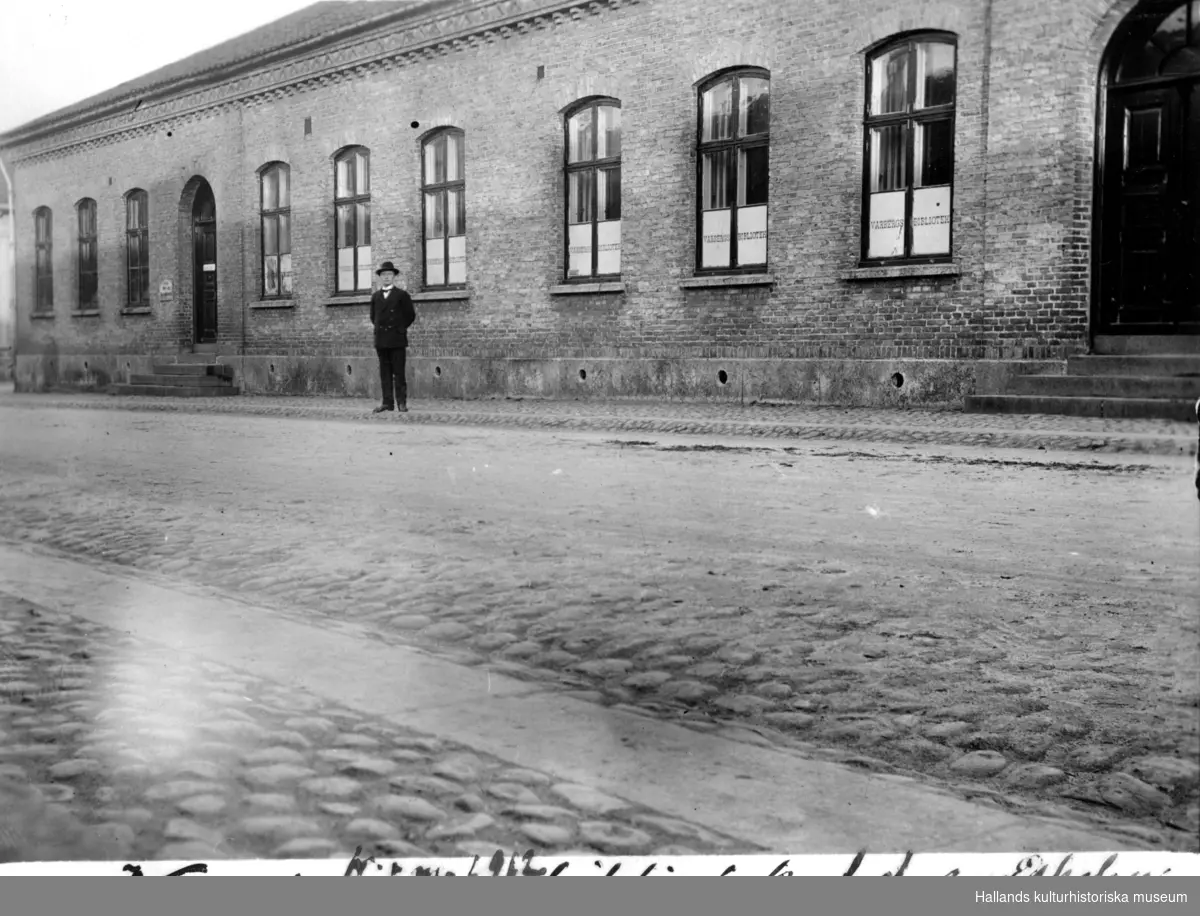 Dåvarande Varbergs bibliotek. Biblioteket flyttade in i lokalerna 1912. Tidigare tjänstgjorde lokalerna som folkskola (enligt anteckning på fotot "f.d. småskolan"). Bilden tagen utmed Östra långgatan, mot nordost, och en man står på trottoaren. Fototid: 1910-tal.