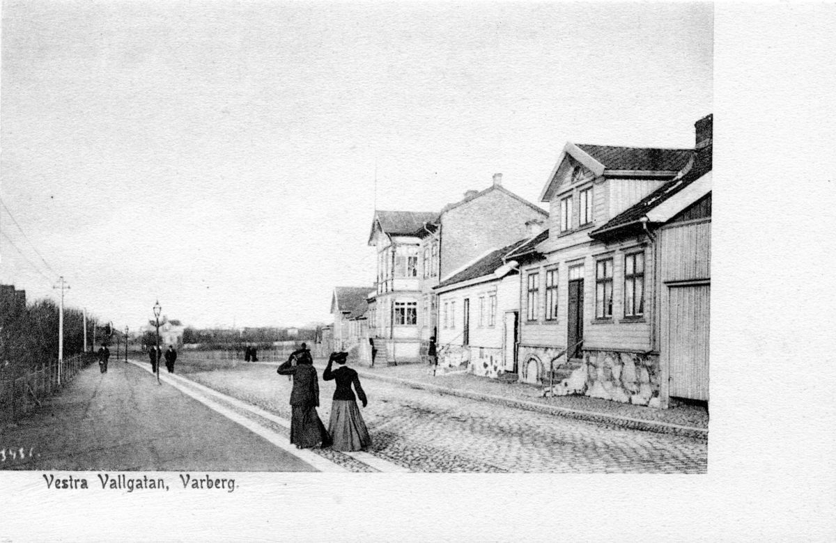 Brevkort, "Vestra Vallgatan, Varberg" (meddelandet skrivs på den tomma ytan på framsidan). Bilden är tagen mot norr så till höger syns kvarteret Berget och längs vänster sida ligger Järnvägsparken. Två damer promenerar och håller i hattarna. De vita stolparna till höger är Hugo Gerlachs elstolpar (uppfördes 1892) som går från hans Mekaniska verkstad, via Järnvägs- och Engelska parkerna till hans bostad i Franckalyckan. Stadens gatlyktor står på gångstråket.