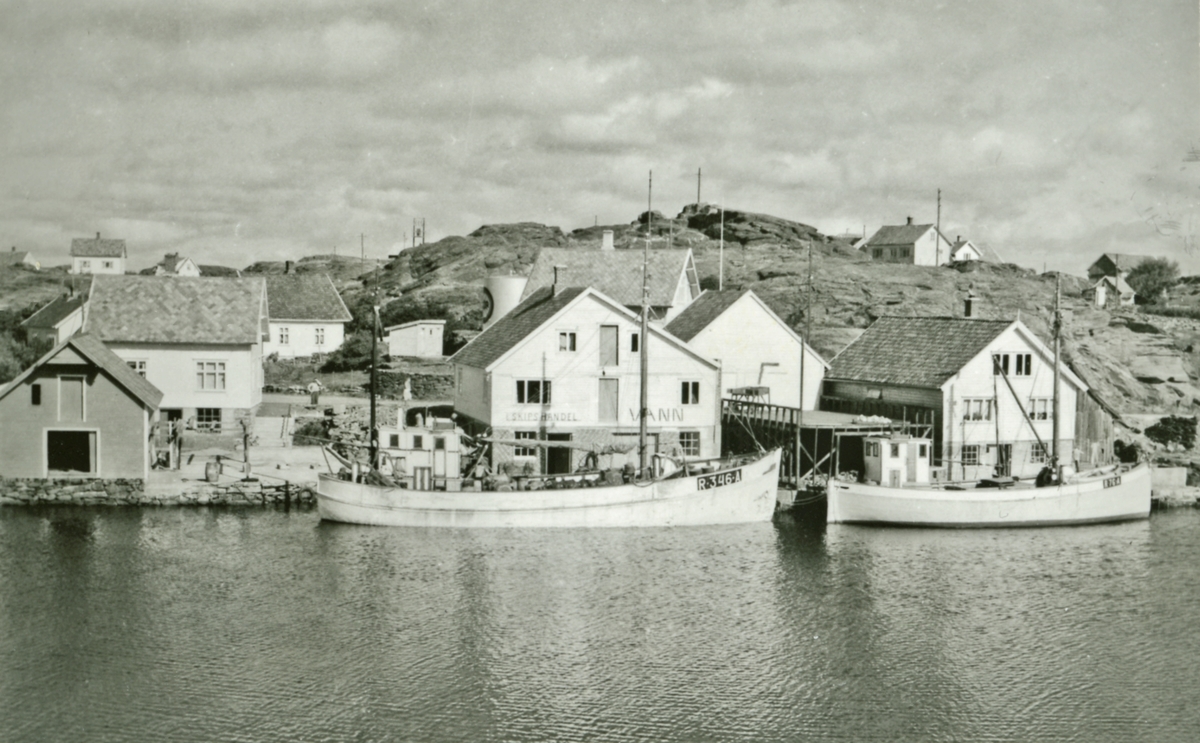 M/S "Solfrid" og M/S "Heim I" ved kai i Sevland. Sjøhus og forretninger på kaien. Bebyggelse og landskap i bakgrunnen.