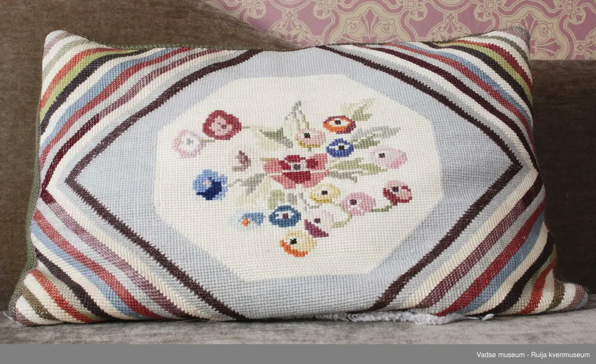 1 stk pute. Tidl. reg. brodert korssting på tysk sekk.Fra ca. 1942-43
Blomstermotiv i midten, striper diagonalt i hjørnene. Strikket grønn bakside.