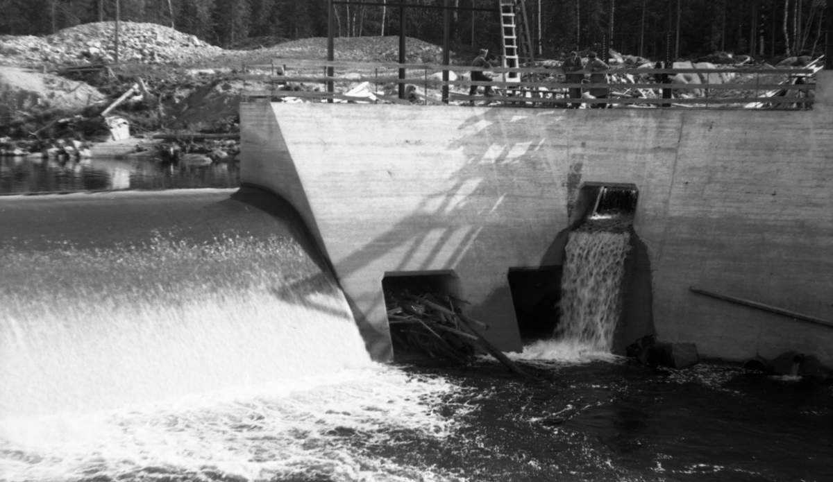 Dammen ved Kvernfallet i elva Søndre Osa i Åmot i Hedmark, fotografert i motstrøms retning i 1941, På dette tidspunktet var dette anlegget, som ble satt i drift i 1936, ennå forholdsvis nytt, og i bakgrunnen på bildet ser vi fortsatt jord- og grushauger fra anleggsperioden. Hovedmotivet her er imidlertid damkonstruksjonen, som var utført i armert betong. Til venstre i bildet ser vi hvordan vannet rant over overløpsdelen, som hadde avrundet krone. I midten og til høyre på bildet ser vi en mer kantete mur som omsluttet det bassenget der turbinrøret hadde sitt innløp. Det skulle føre driftsvann ned til kraftstasjonen ved Brufallet, noen hundrede meter lengre nede ved elveløpet. Turbinrøret vises ikke på dette fotografiet, men vi ser tre noenlunde kvadratiske åpninger i dammuren, to helt nederst og en litt høyere oppe. Gjennom den sistnevnte strømmet det vann da dette bildet ble tatt. Disse åpningene skulle antakelig brukes som avløpsventiler når det var vel mye vann i dammen. De kunne åpnes og lukkes ved hjelp av hev- og senkbare luker på et stålstativ som var montert på motstrøms side av dammuren. Da dette fotografiet ble tatt sto det tre karer og en stige på damkrona.