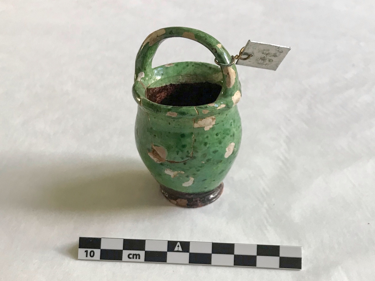 Liten grønn leirkrukke med handtak. Glasert. 

Har flere revner, delvist ødelagt. Har vært limet sammen.