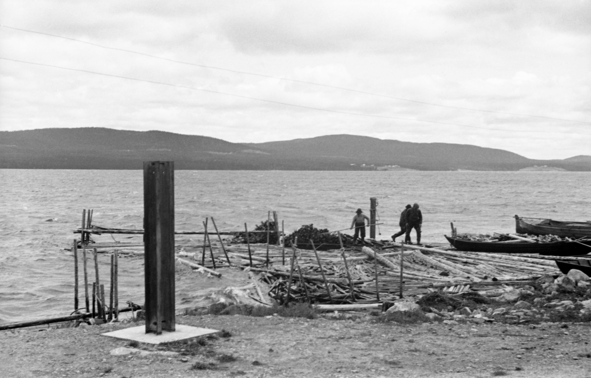 Fra Sørlistøa ved Osensjøen i Åmot.  Fra 1939 hadde Glomma fellesfløtingsforening et administrasjonssenter for fløtinga på sjøen her, med kontor for fløtersjefen, mannskapshus, båthus, vedhus og smie.  Da dette fotografiet ble tatt hadde det vært storm slik at flåten med ved hadde gått istykker.  Bildet viser tre karer som arbeider med oppsamling av ved i området ved brygga.  På stranda til høyre ligger et par robåter.  I forgrunnen til venstre står et par vertikale stålstenger nedstøpt i ei betongkube, en konstruksjon som antakelig er bygd som flaggstangfundament. 