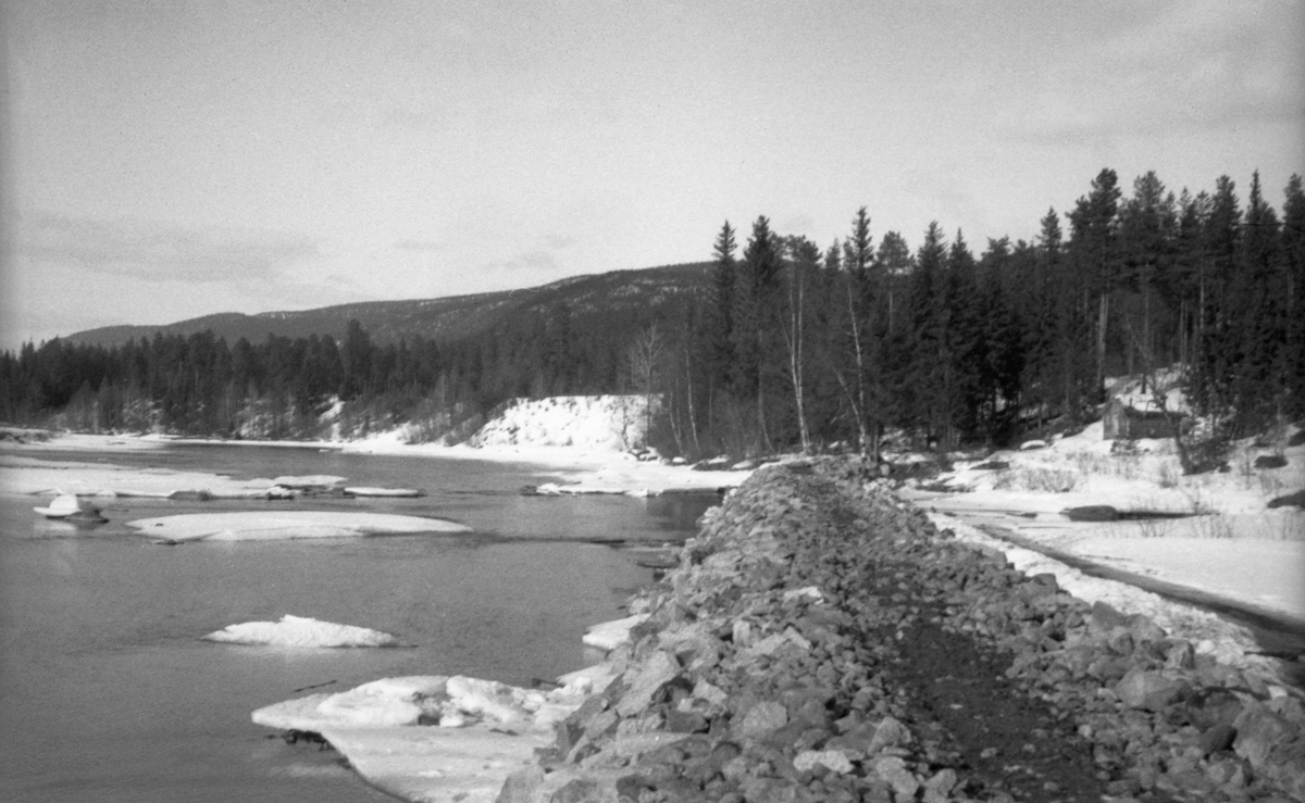 Skådam av stein i ved Søråsenget i Søndre Osa i Åmot kommune i Hedmark. Denne skådammen er en voll av stein og grus, sannsynligvis hentet fra elveløpet og skjøvet sammen ved hjelp av en bulldoser. Skådammen skulle hindre at fløtingsvirke drev inn i strandsona og ble liggende der, slik at fløterne fikk en besværlig jobb med å løsne det og få det til å flyte videre. Til høyre i bildet ser vi et lite hus, antakelig ei koie. Fotografiet ble tatt i slutten av mars 1946, på et tidspunkt da det fortsatt lå snø i terrenget langs vassdraget.