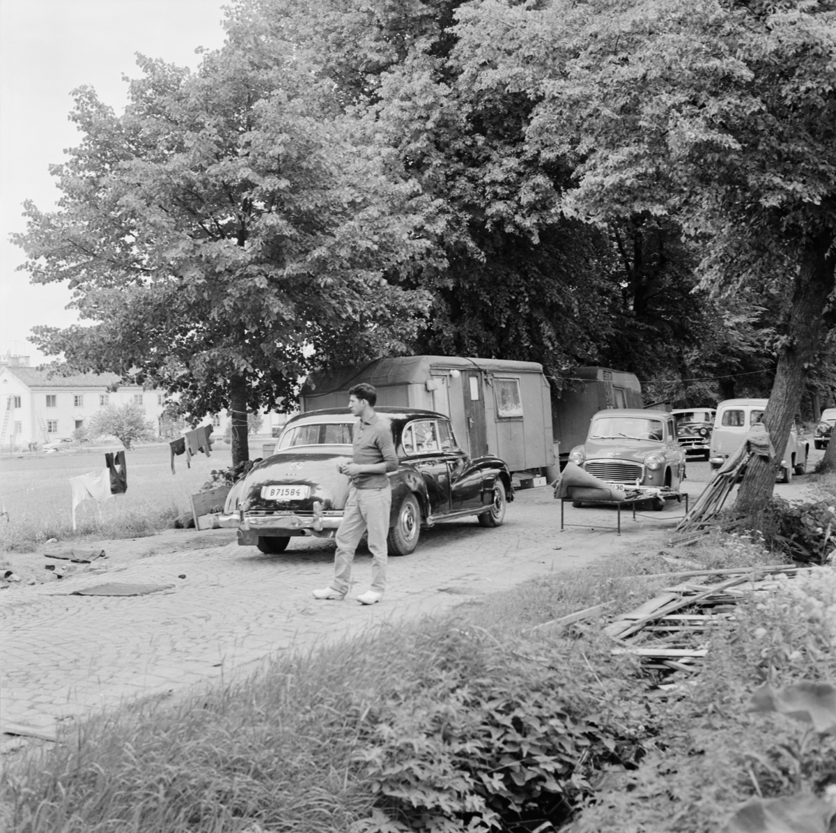 En man står på gatan invid husvagnar och bilar, Uppsala, juli 1963