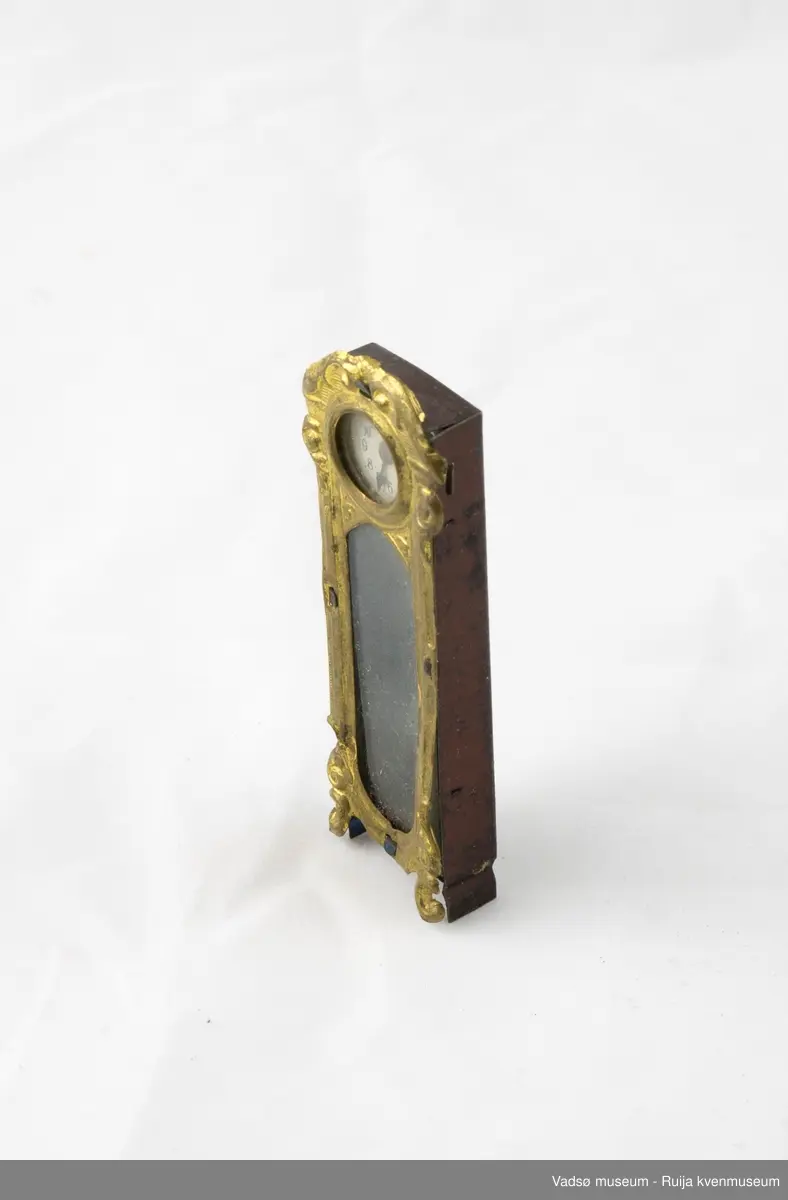Dukkemøbel. Gulvur i miniatyrstørrelse. Kasse i brunlakkert metall. Gullfarget frontpanel med to vinduer; ett til tallskive, og ett til pendel. Opptrekksfunksjon på baksiden.