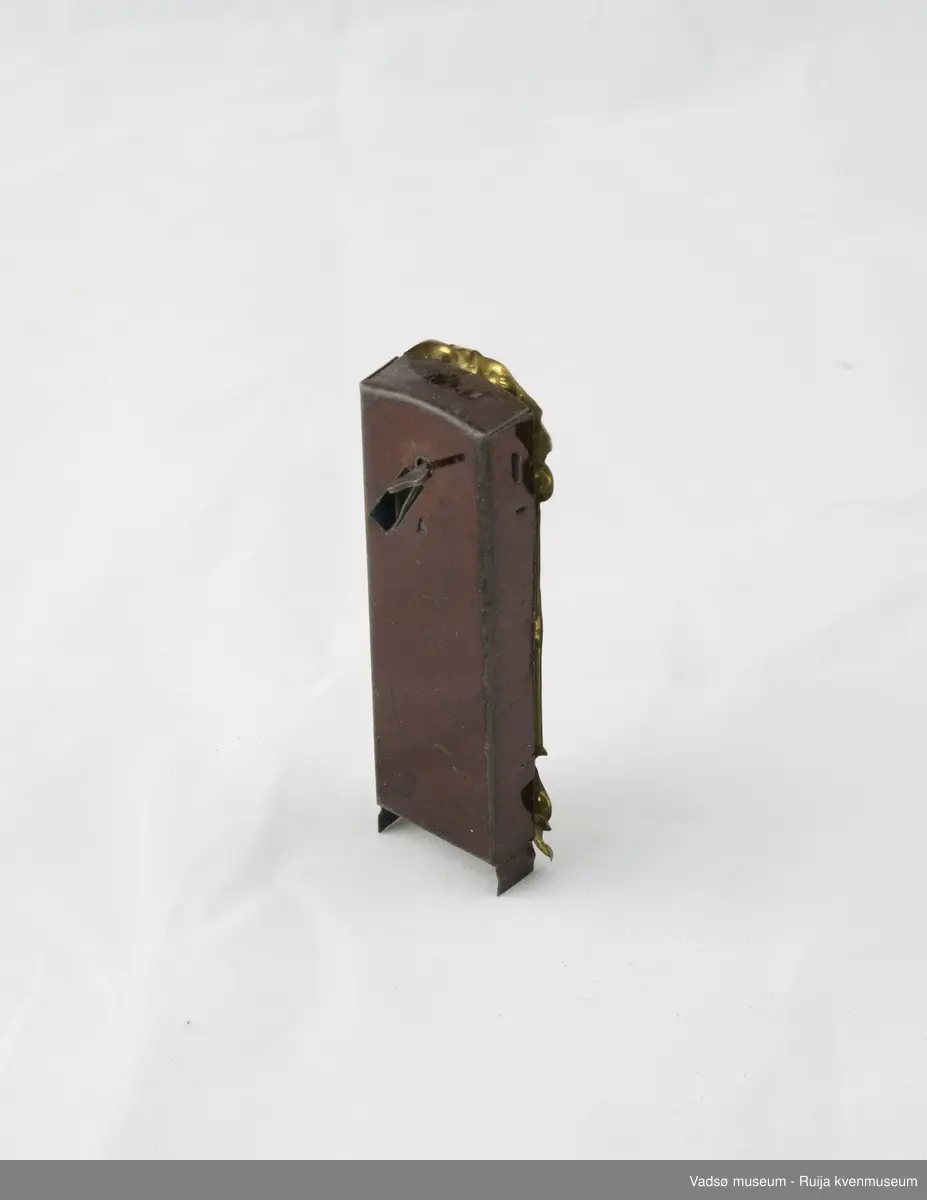 Dukkemøbel. Gulvur i miniatyrstørrelse. Kasse i brunlakkert metall. Gullfarget frontpanel med to vinduer; ett til tallskive, og ett til pendel. Opptrekksfunksjon på baksiden.