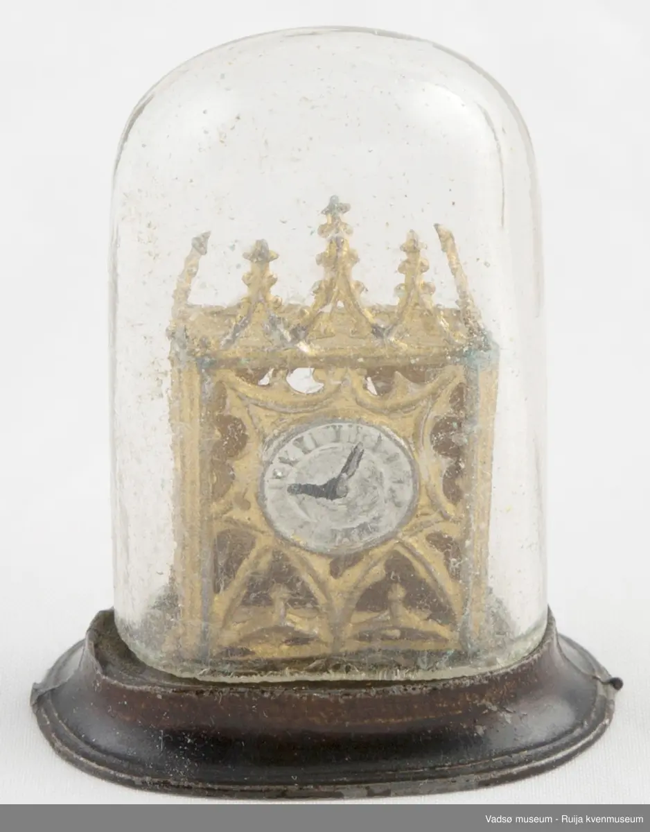 Dukkehusinventar. Forestiller bordklokke i metall. Rektangulært klokkehus med 5 spir i front. Klokkehus er loddet fast i oval metallsokkel som er noe bredere enn klokkehuset. Urskive i sølv og visere i svart. Klokka har en pynte/beskyttelseskuppel i tykt glass som kan tas av og på.