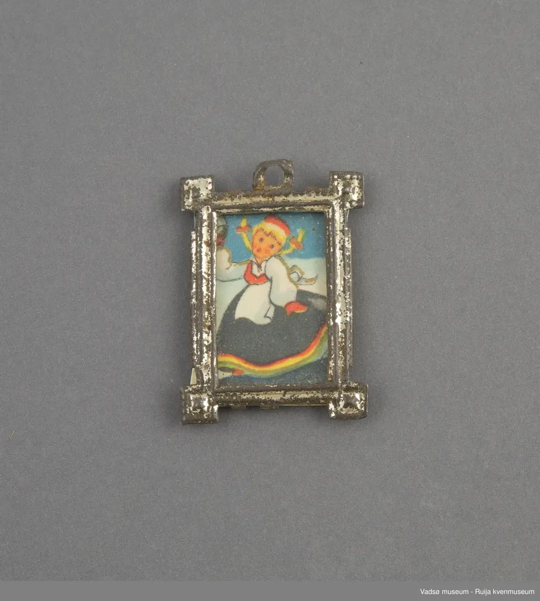 Dukkehusinventar. Bilde med glass og ramme. Rektangulær ramme i sølvfarget tynt metall, utformet i art deco stil. Kvadratisk vulst/ornament i hvert hjørne, øre med rundt opphengshull midt på ene kortsiden. Glasset er festet fast i rammen på baksiden.