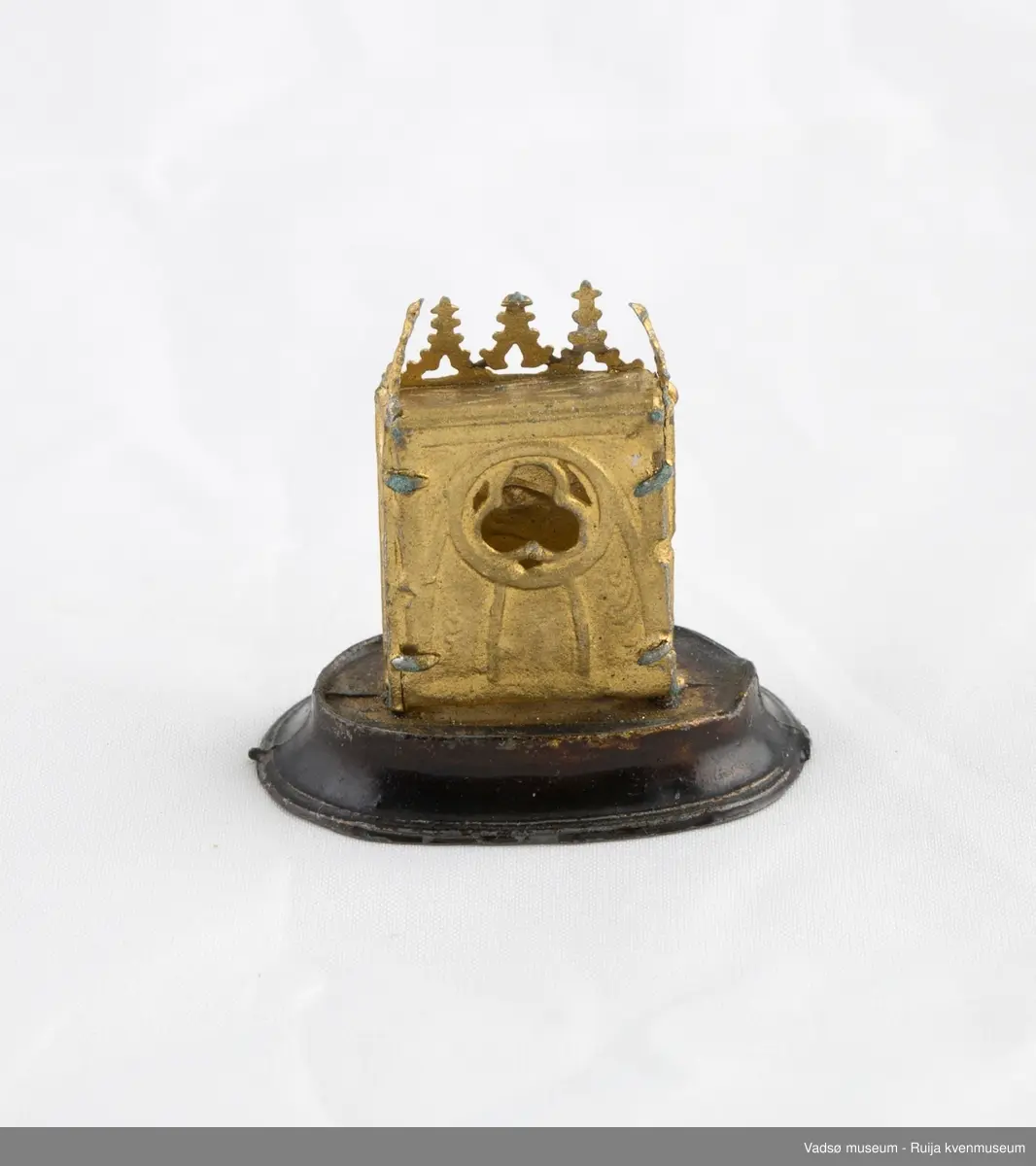 Dukkehusinventar. Forestiller bordklokke i metall. Rektangulært klokkehus med 5 spir i front. Klokkehus er loddet fast i oval metallsokkel som er noe bredere enn klokkehuset. Urskive i sølv og visere i svart. Klokka har en pynte/beskyttelseskuppel i tykt glass som kan tas av og på.