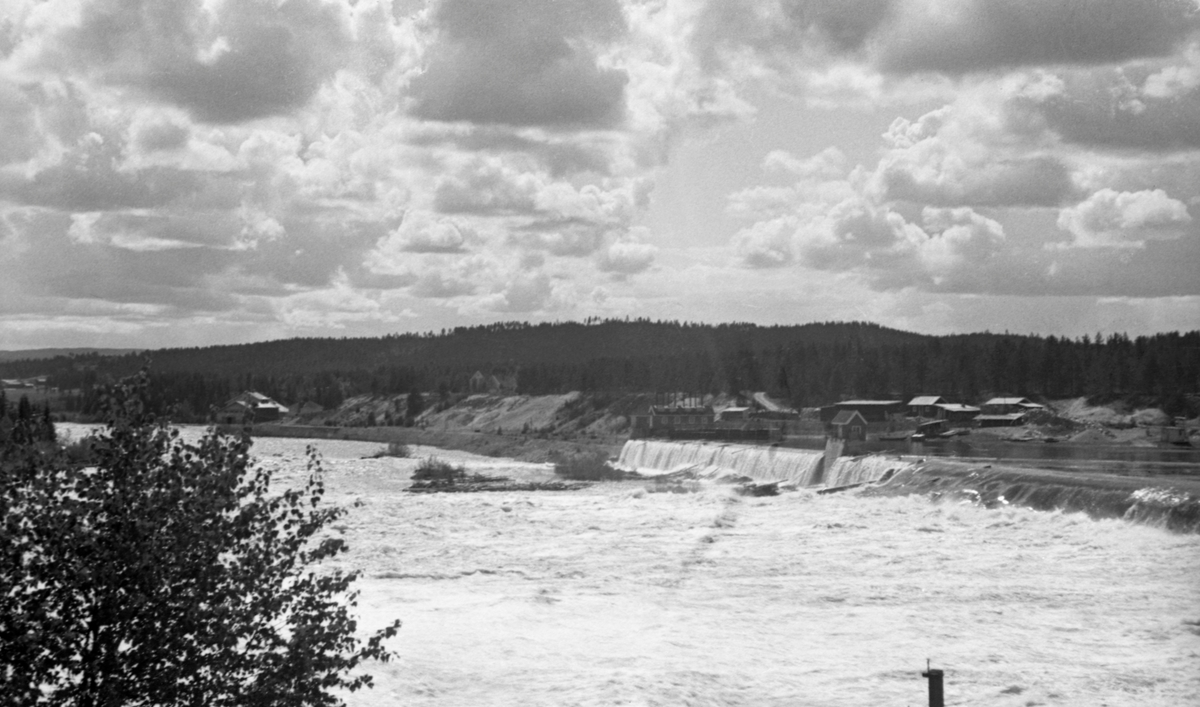 Skjefstadfossen i Glomma i Heradsbygda i Elverum, fotografert en forsommerdag i 1944 da vannføringa i elva var stor. Her ble det første store kraftverket i Glomma bygd i 1909-1910.  I samband med dette prosjektet ble det støpt en drøyt 300 meter lang terskeldam på tvers av elveløpet. Denne konstruksjonen skulle stuve opp vann i den ovenforliggende delen av elveløpet og sørge for kontinuerlig tilsig til kraftverksdammen i den vestre delen av elveløpet, som vi ser til venstre i dette bildet. Terskeldammen ble gjort noe lavere mot den østre elvebredden. Poenget med dette var å skape et strømdrag der som trakk mesteparten av fløtingstømmeret mot den østre delen av elveleiet, som var djupere enn midtartiet, der det hadde lett for å bygge seg opp digre hauger med fløtingstømmer.  Da dette fotografiet ble tatt lå det litt tømmer under den midtre delen av dammen, men ellers var vannføringa stor nok  til at det meste ble skylt over steinørene like nedenfor den nevnte terskelen i elveløpet.