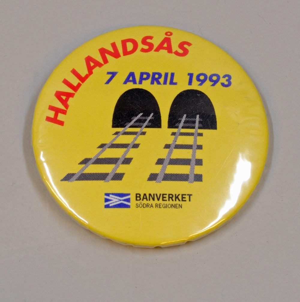 Gul reklamknapp med texten "HALLANDSÅS 7 APRIL 1993", texten är tryckt i orange och blått. I mitten finns en bild av två spår som går in i varsin tunnelöppning. Längst ned finns logotypen för Banverket samt texten "BANVERKET SÖDRA REGIONEN".