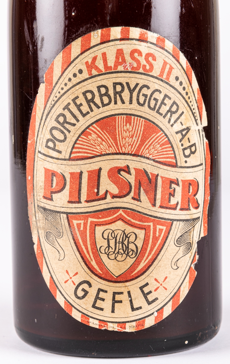 Pilsnerflaska med innehåll. Brunt glas. Etikett: Klass II pilsner, Porterbryggeri AB Gefle.